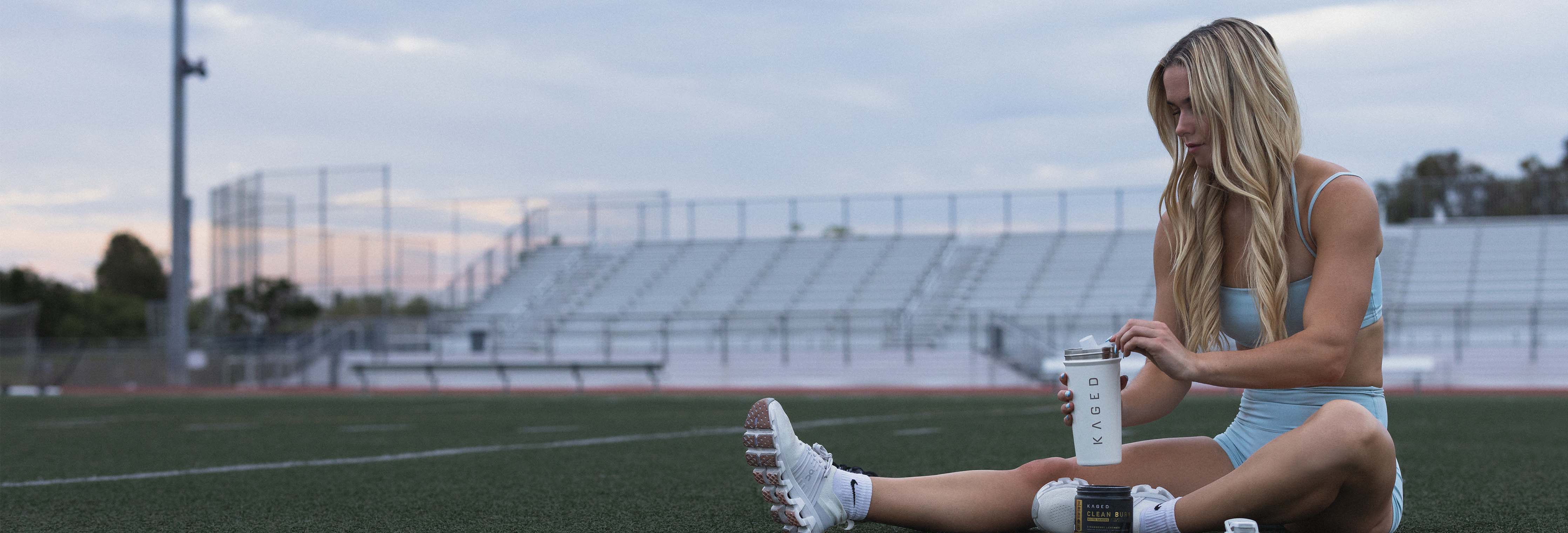 Female athlete sitting on turf stretching and opening a KAGED shaker with supplement containers beside her on a track at sunset
