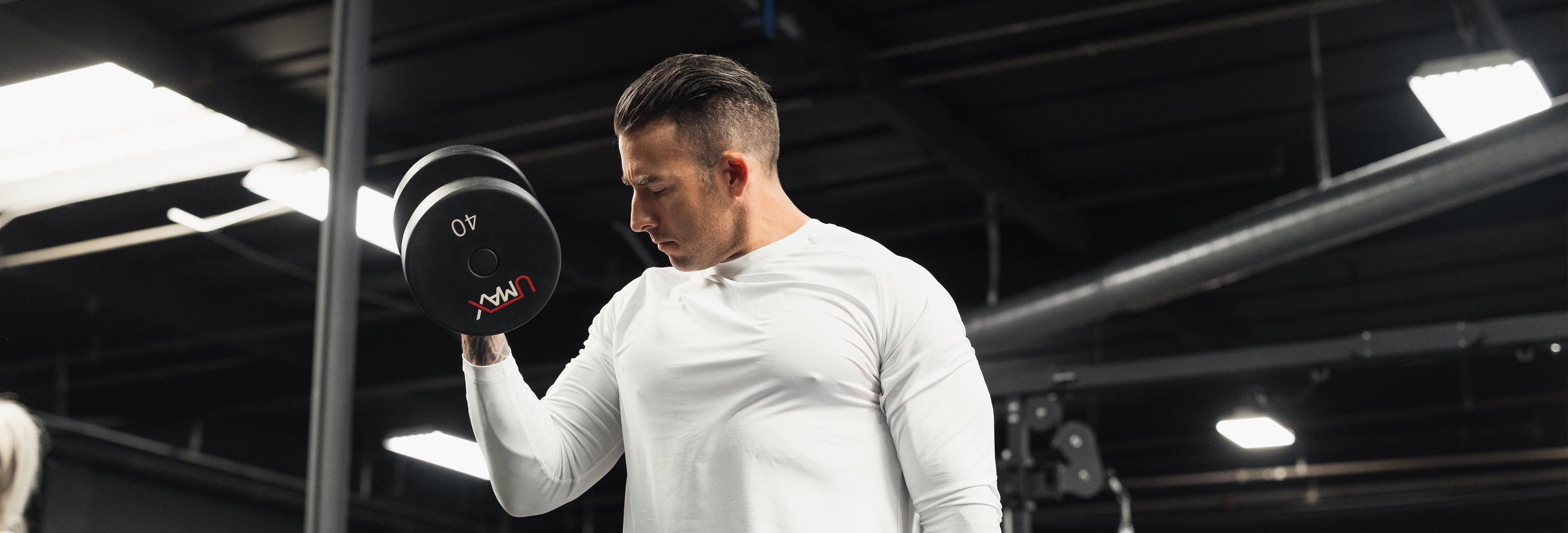 Man performing a standing dumbbell biceps curl in a commercial gym with black ceiling and weight racks in background