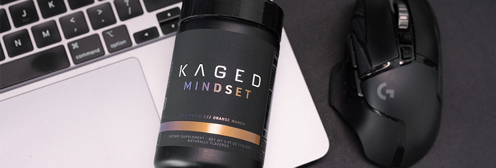 Angled front view of KAGED Mindset supplement tub, Orange Mango flavor, next to laptop and mouse on dark desk