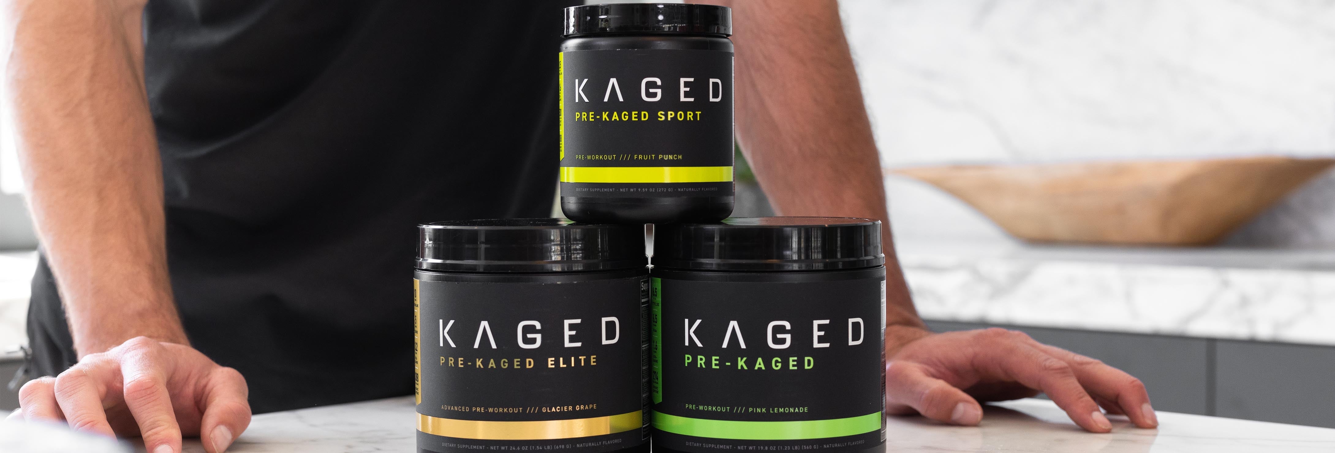 Front view of three KAGED pre-workout tubs stacked showing brand labels and colored label bands on a marble countertop with a person’s hands behind