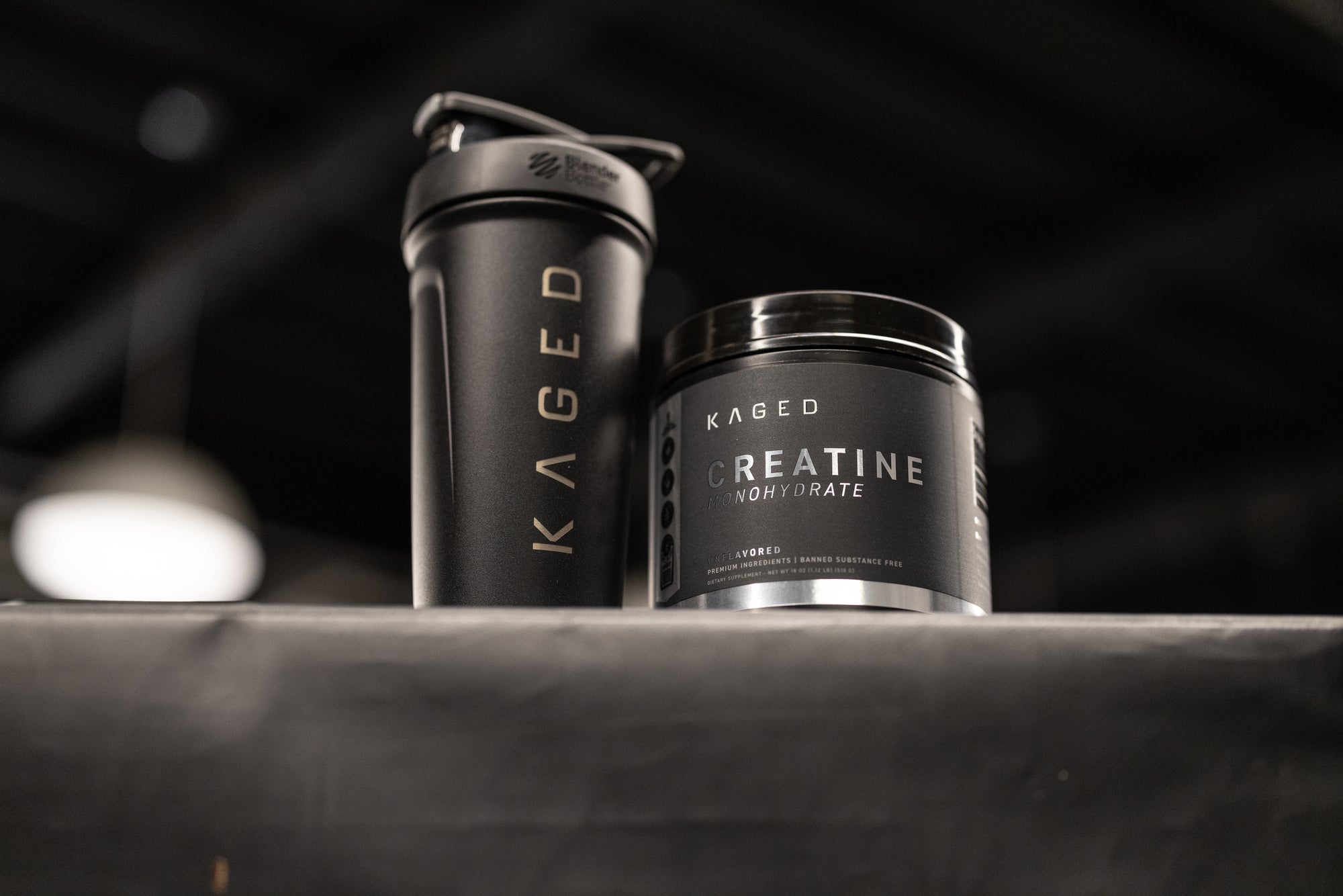 Front view of KAGED Creatine Monohydrate tub beside a black KAGED shaker showcasing product label and brand logo on a dim gym background
