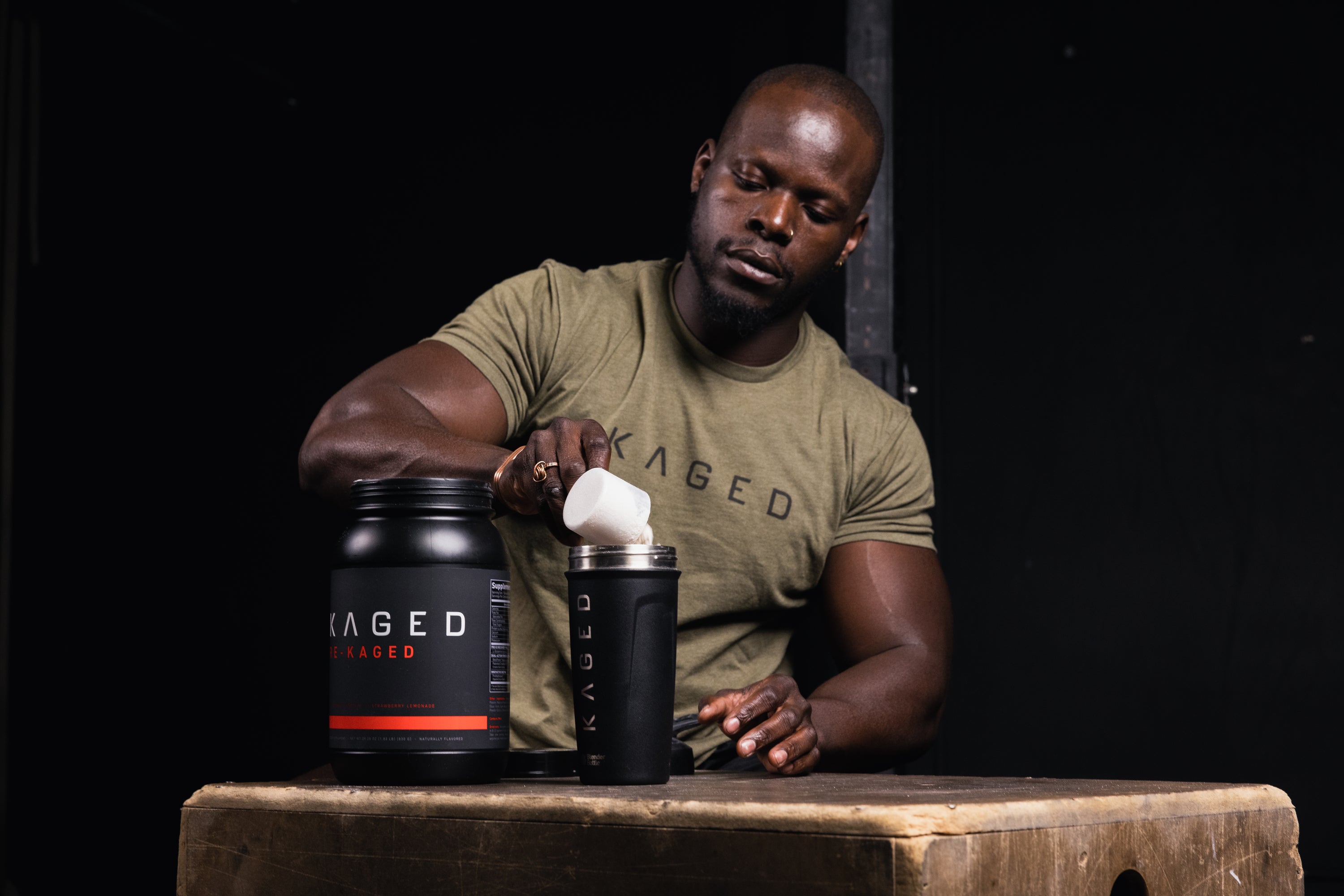 Man scooping supplement powder into a KAGED-branded shaker next to a KAGED protein tub on a wooden box