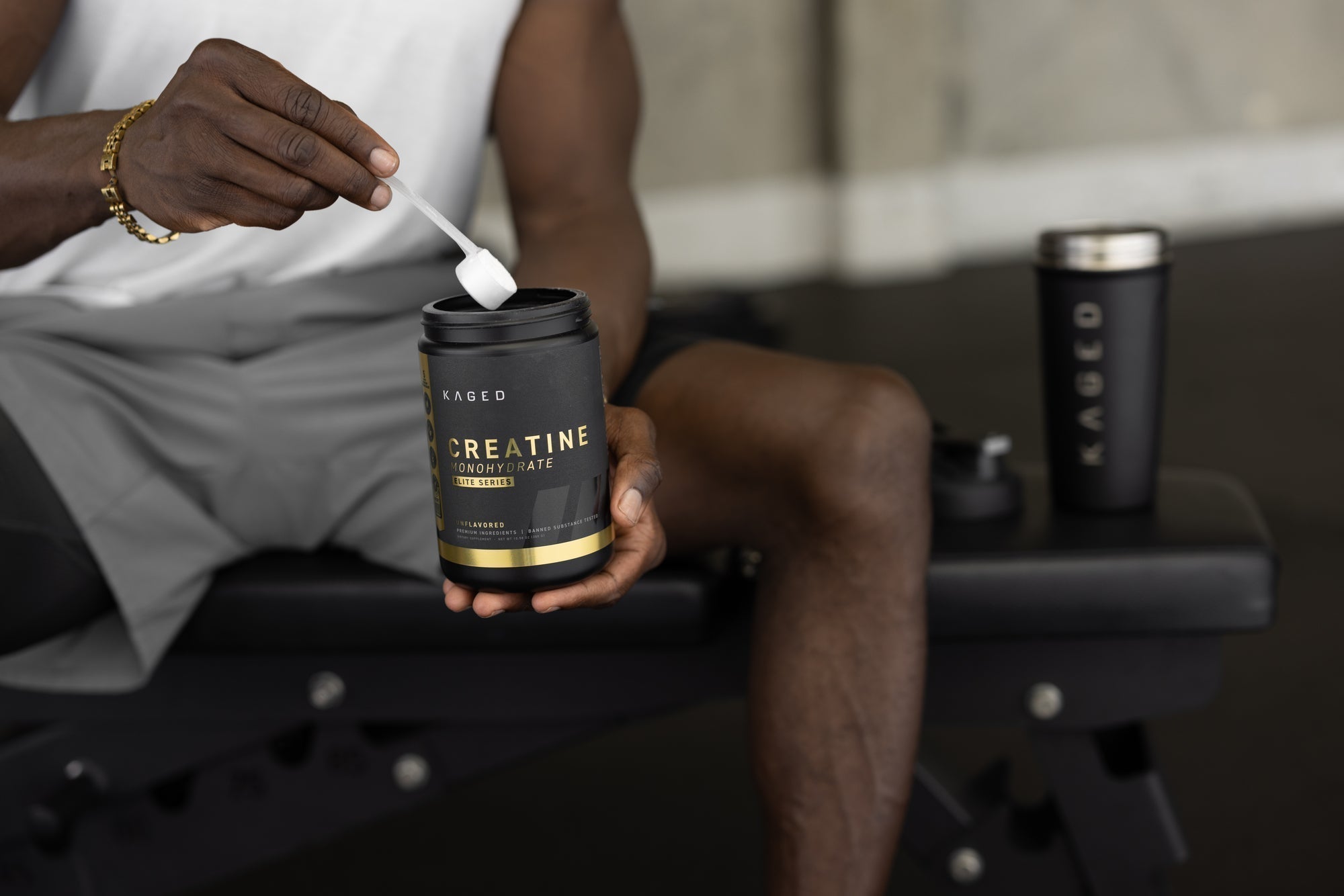 Person scooping Kaged Creatine Monohydrate from black tub while seated on a gym bench, Kaged shaker beside them