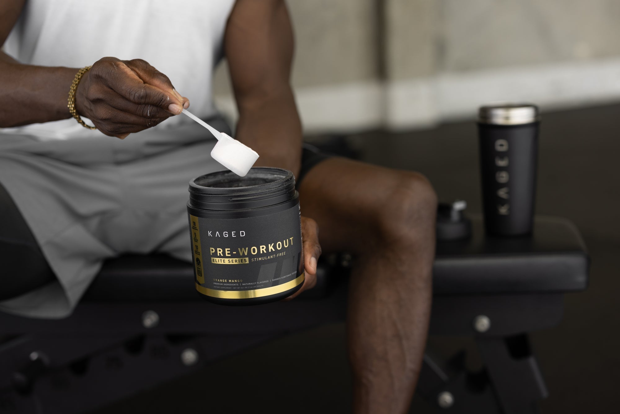 Person scooping KAGED Pre-Workout Elite Series powder from a black tub while seated on a gym bench with shaker nearby