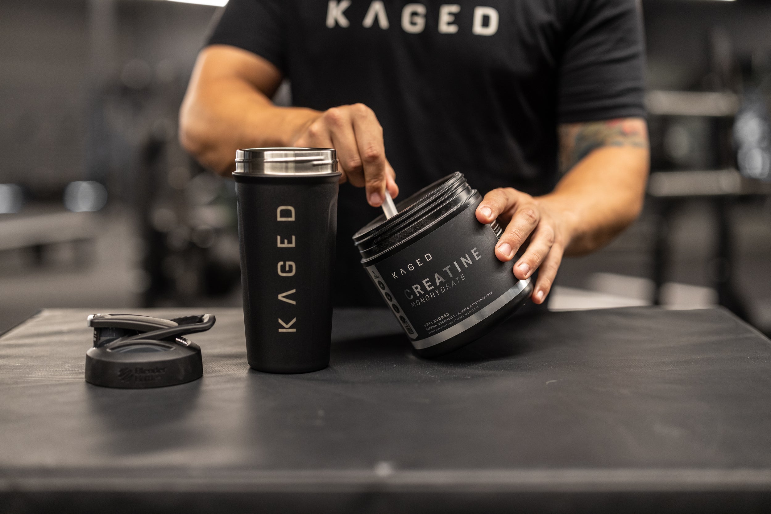 Close up of KAGED Creatine Monohydrate tub being scooped next to a KAGED shaker on a gym bench