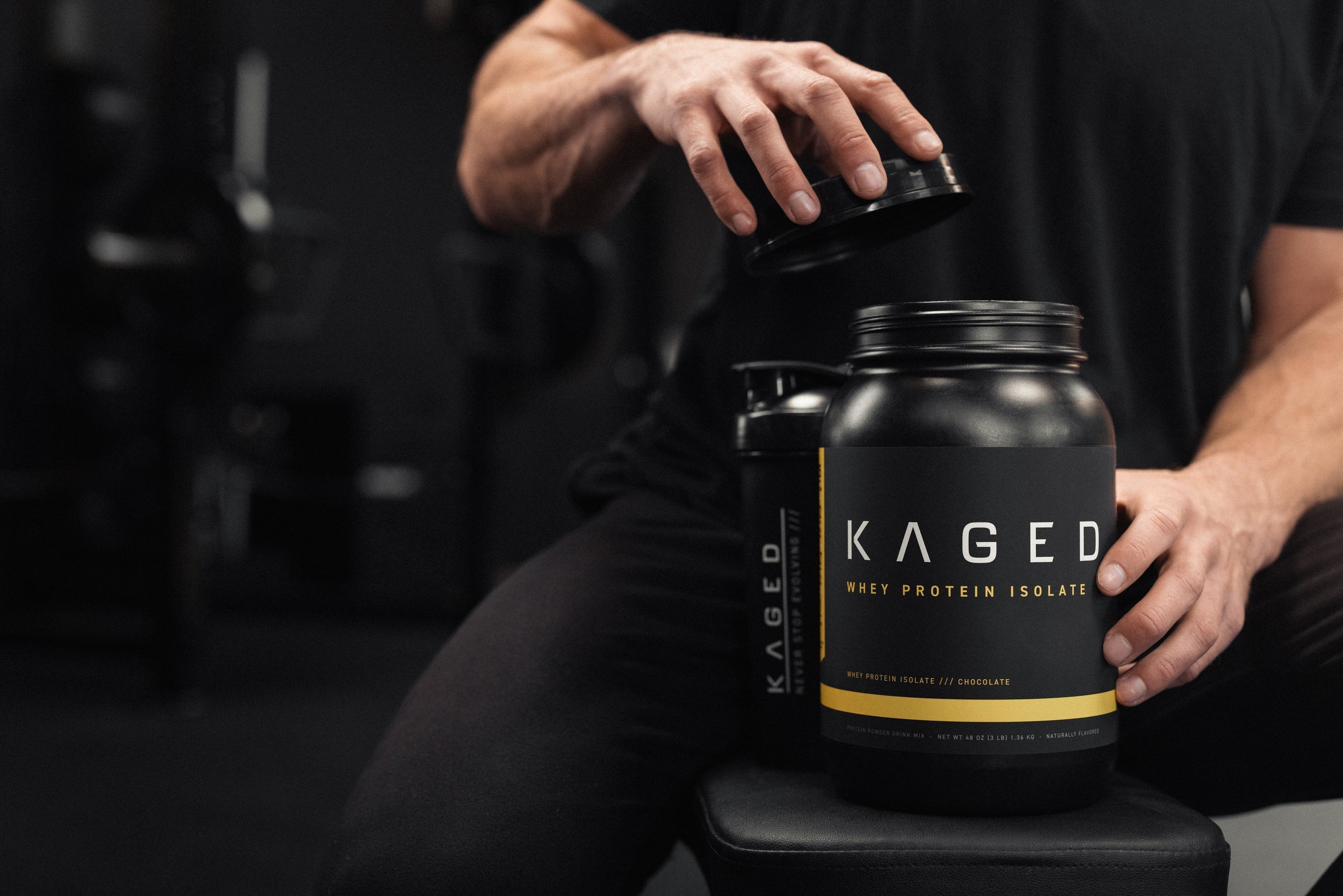 Front view of Kaged Whey Protein Isolate tub showcasing the black label and yellow accent, held on a gym bench with a shaker bottle and blurred gym equipment in background