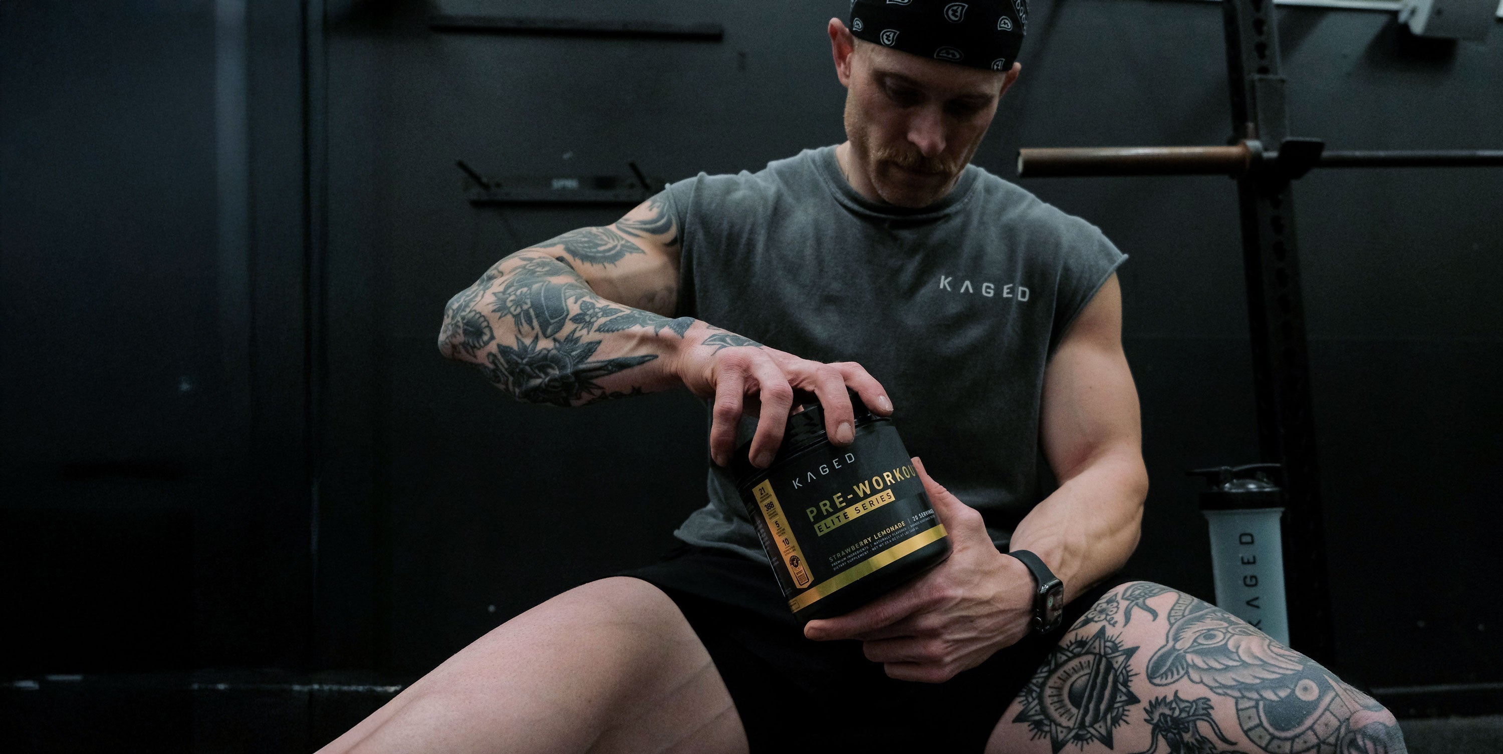 Man in gym opening a KAGED pre-workout tub while seated on a bench, tattoos visible and a KAGED shaker at his side