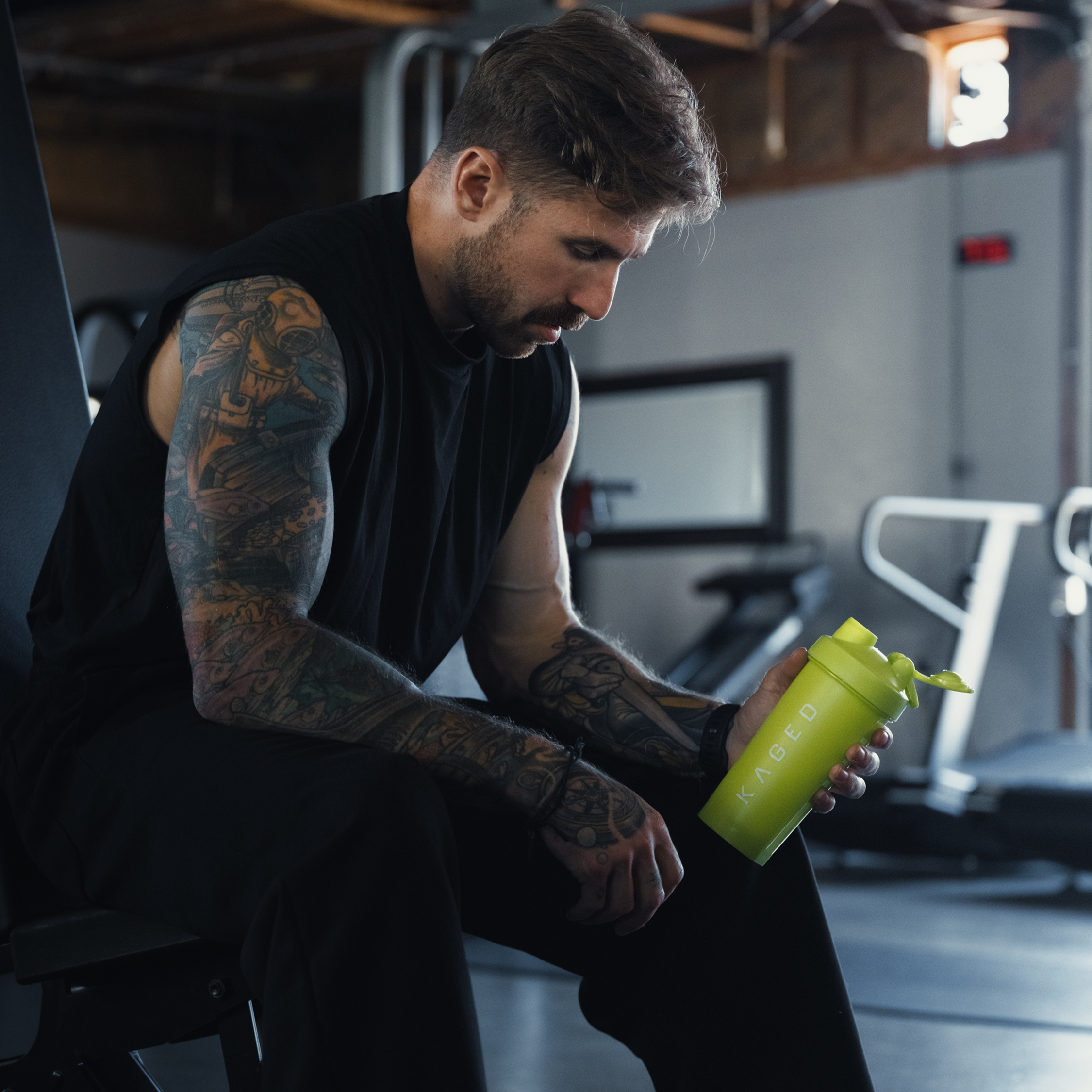 Tattooed man sitting on a gym bench, resting and holding a lime green KAGED shaker bottle