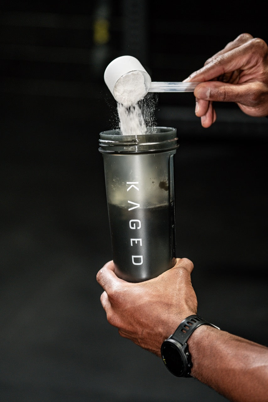 Hand pouring L-Citrulline powder from a scoop into a black KAGED shaker in a gym setting