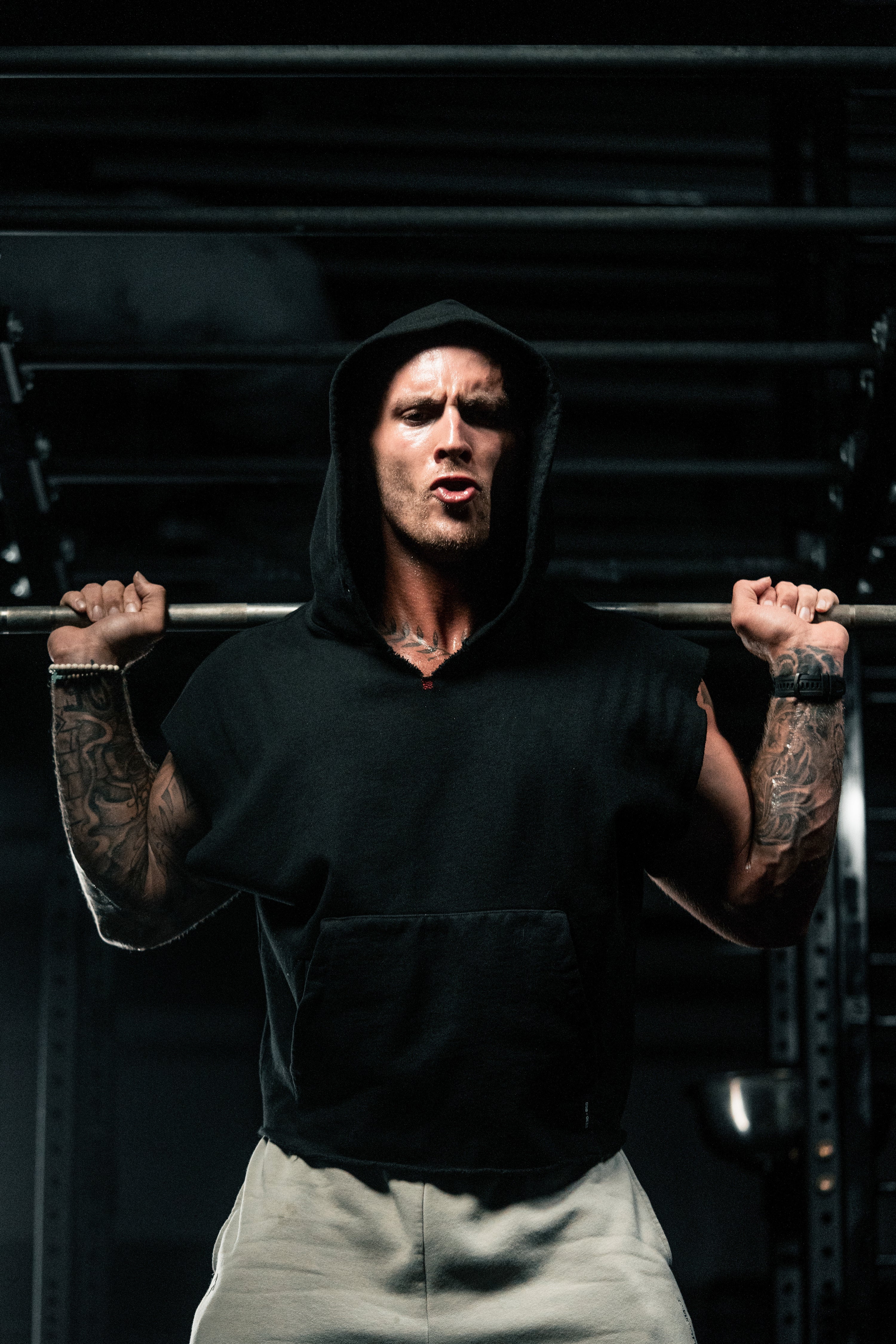 Male athlete wearing a black sleeveless hooded top performing a barbell back squat in a dimly lit gym