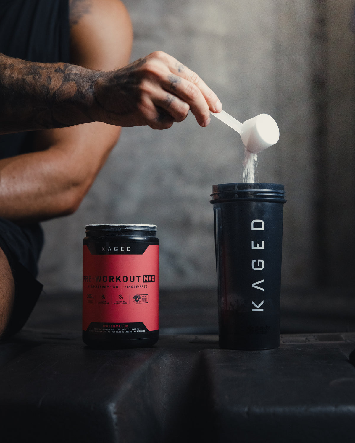 Close-up of Pre-Workout Max tub with scoop pouring powder into a Kaged shaker, watermelon flavor label visible