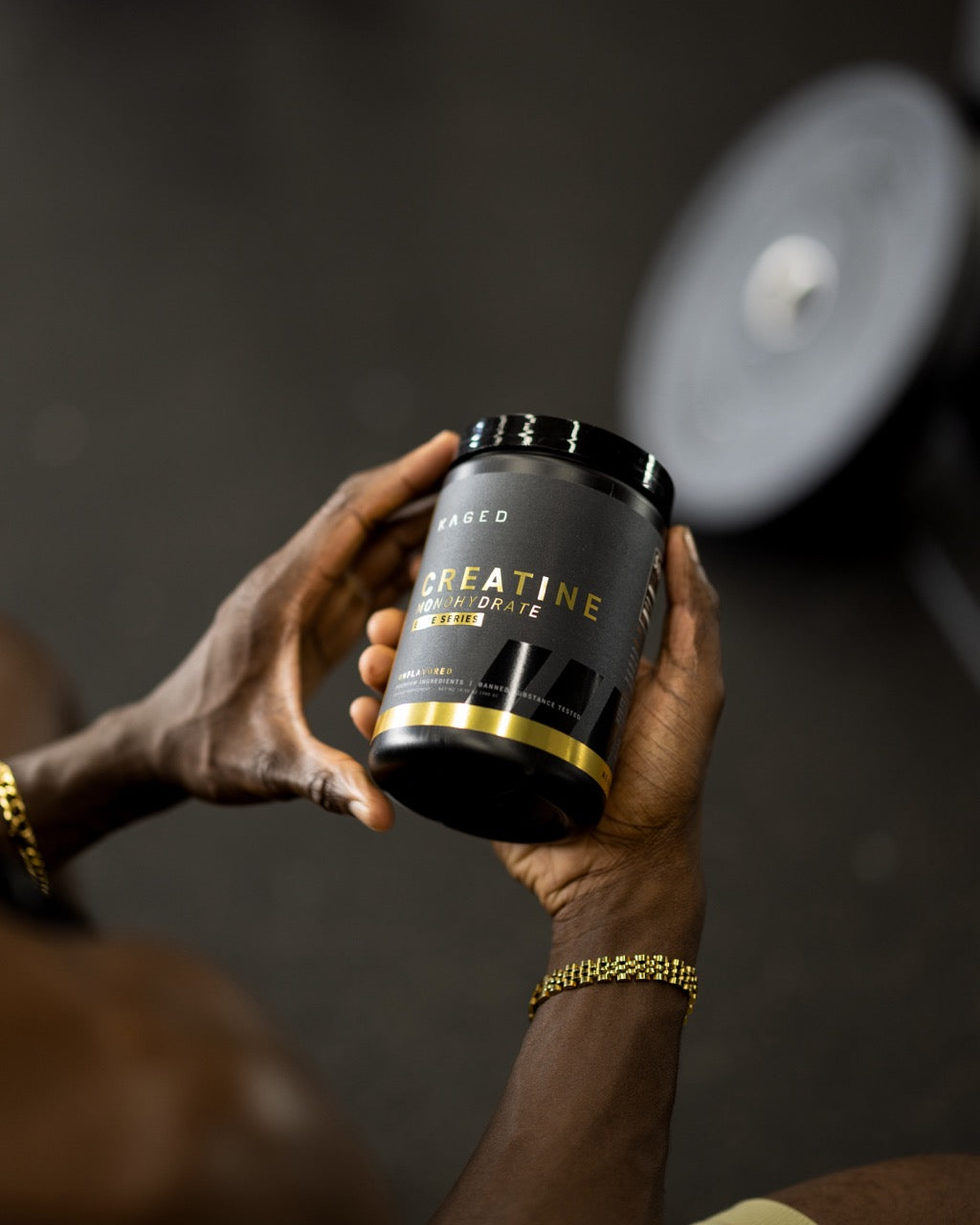 Angled view of KAGED Creatine jar showing CREATINE gold-accent label held by person in gym with blurred weight plate