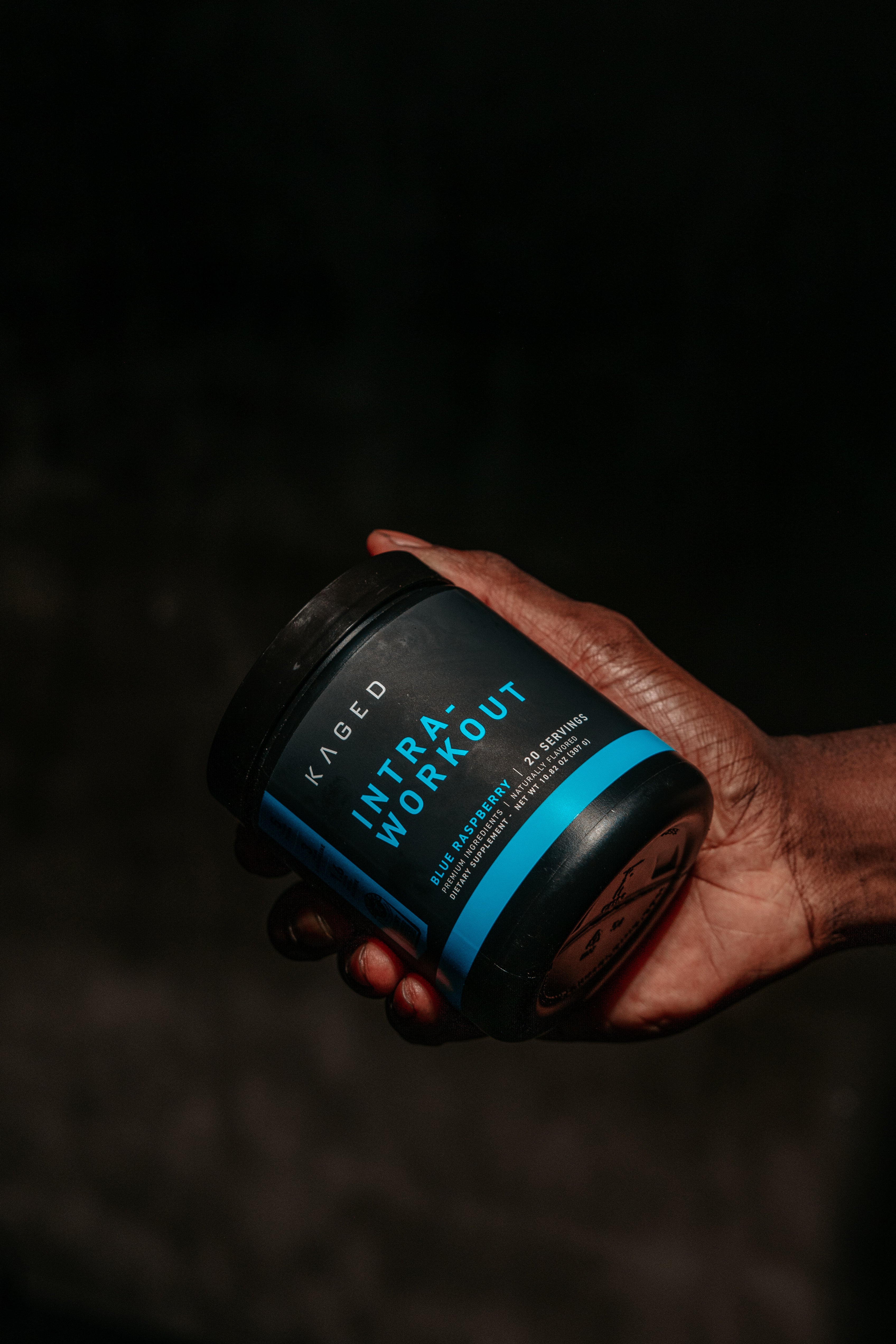 Angled close-up of Intra-Workout jar with blue INTRA-WORKOUT label, Blue Raspberry flavor, held in a hand on dark background