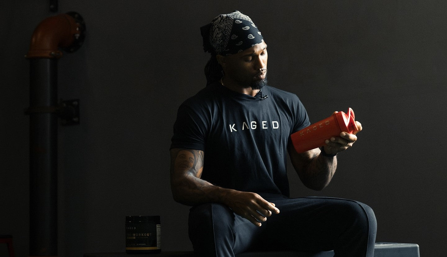 Man wearing KAGED t-shirt inspecting a red KAGED shaker bottle while seated in a dim gym with a supplement tub nearby