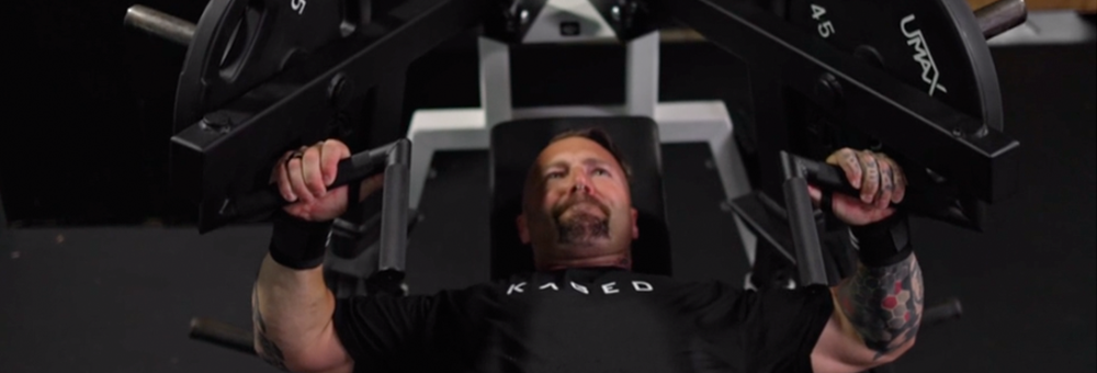 Man performing seated machine shoulder press in a gym setting, wearing a KAGED t-shirt