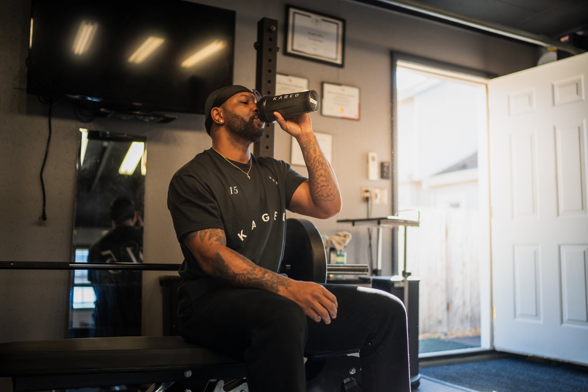Man drinking from a KAGED shaker while seated on a weight bench in a home gym, wearing a KAGED t-shirt