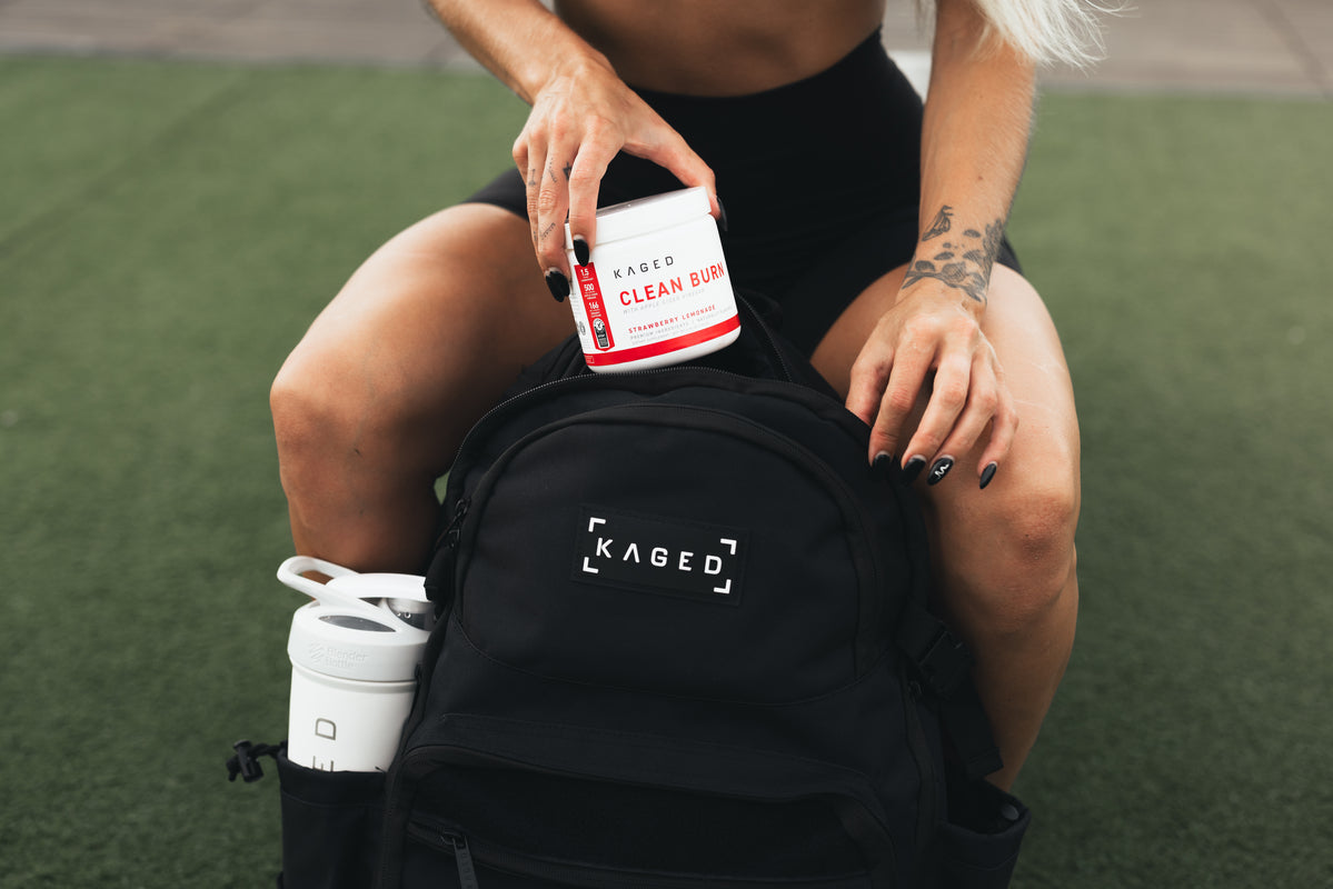Person placing a KAGED Clean Burn supplement tub into a black KAGED backpack on outdoor turf with a white shaker in the bag