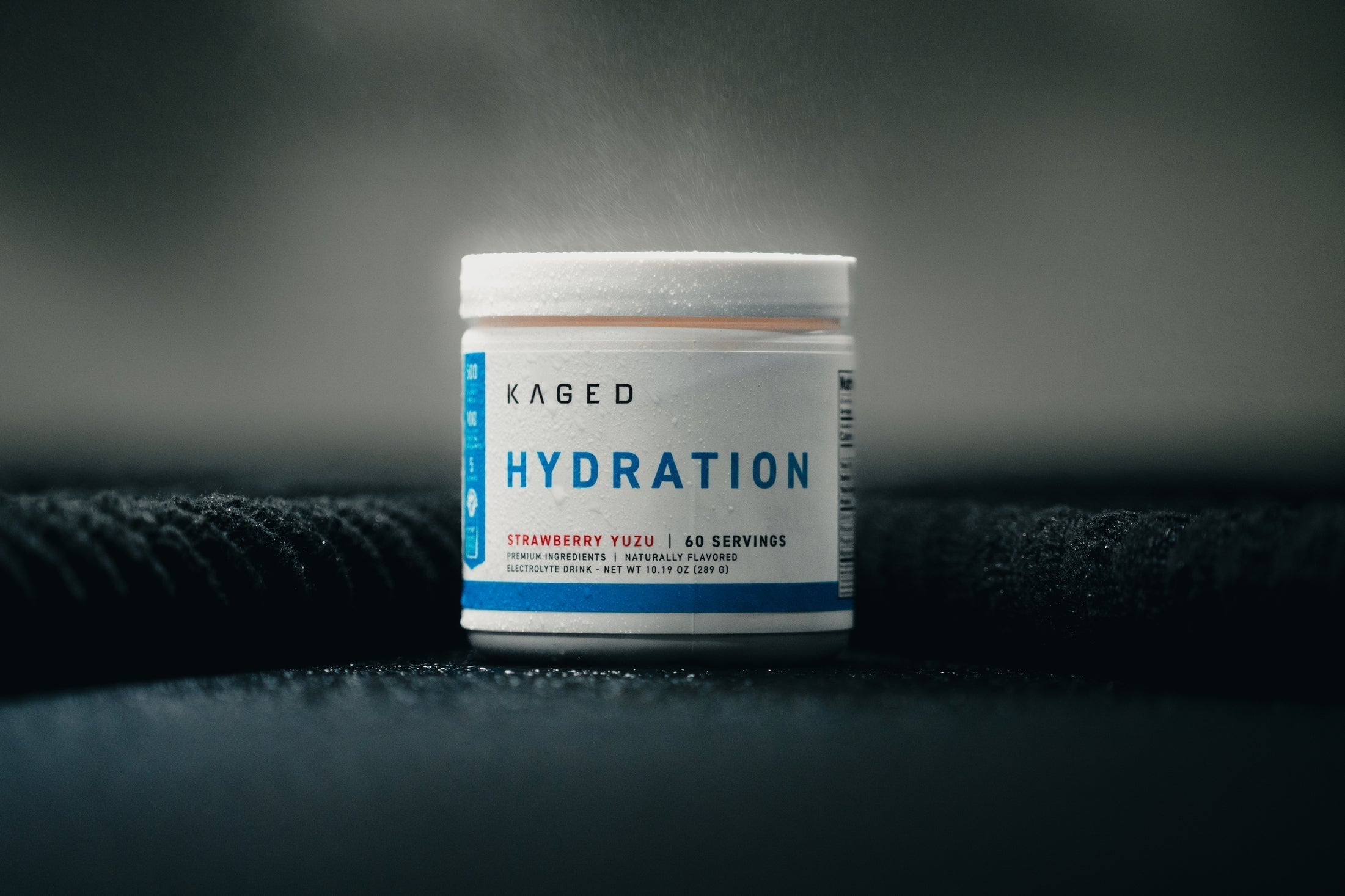 Front view of KAGED Hydration electrolyte powder tub showing Hydration label, Strawberry Yuzu flavor and 60 servings on dark textured background