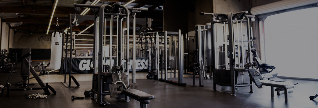 Empty gym interior showing cable machines, benches, and free weights in a dimly lit training space