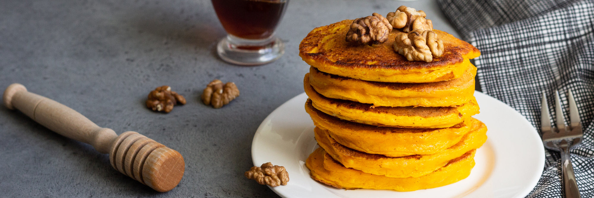 Stack of protein pumpkin pancakes topped with walnuts on a white plate beside a honey dipper, glass of coffee, and fork on a gray surface