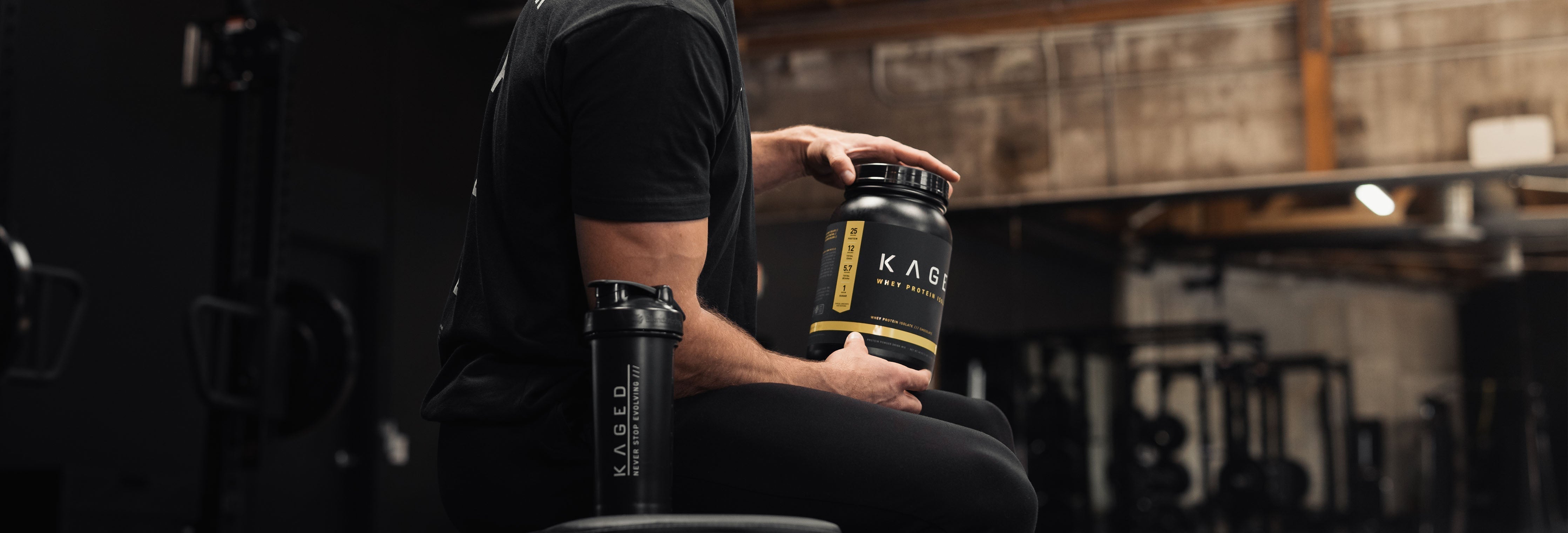 Man seated on a gym bench holding a Kaged whey protein tub with a Kaged shaker beside him