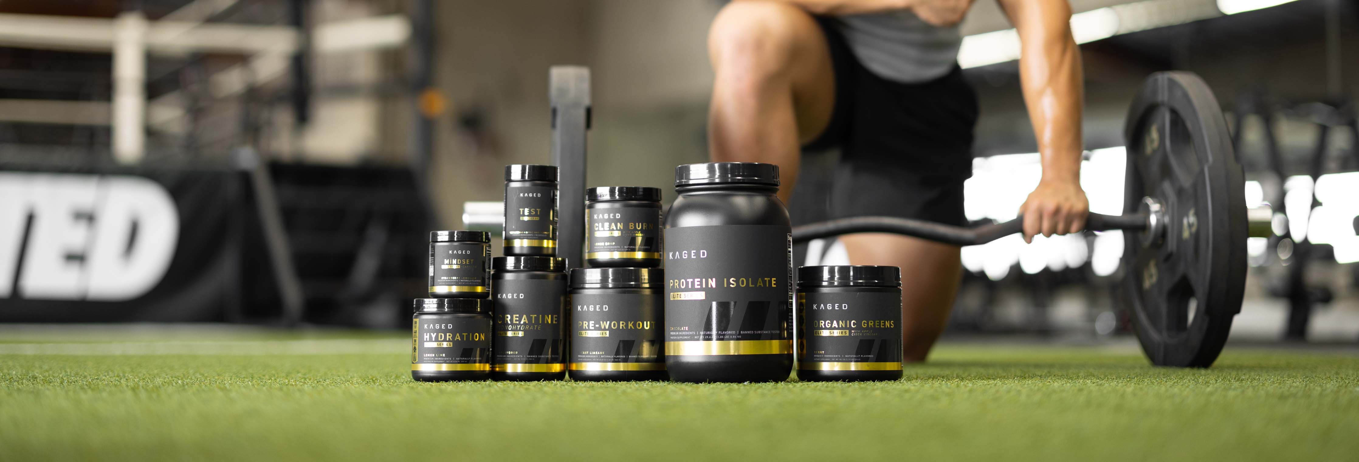 KAGED supplements (protein, creatine, pre-workout) arranged on gym turf with athlete deadlifting in background