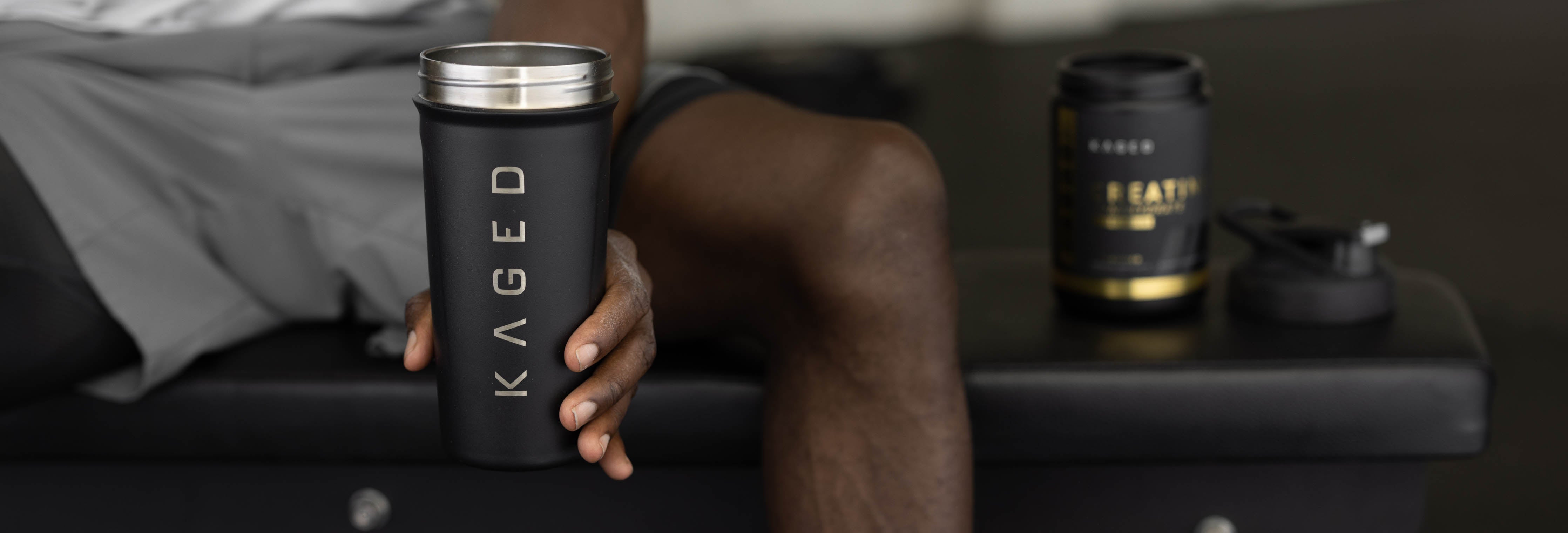 Athlete holding a black KAGED shaker cup on a gym bench with a KAGED supplement tub in the background
