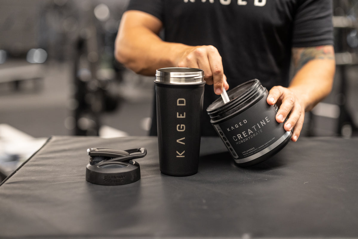 Person scooping Kaged creatine monohydrate powder into a Kaged shaker cup in a gym setting