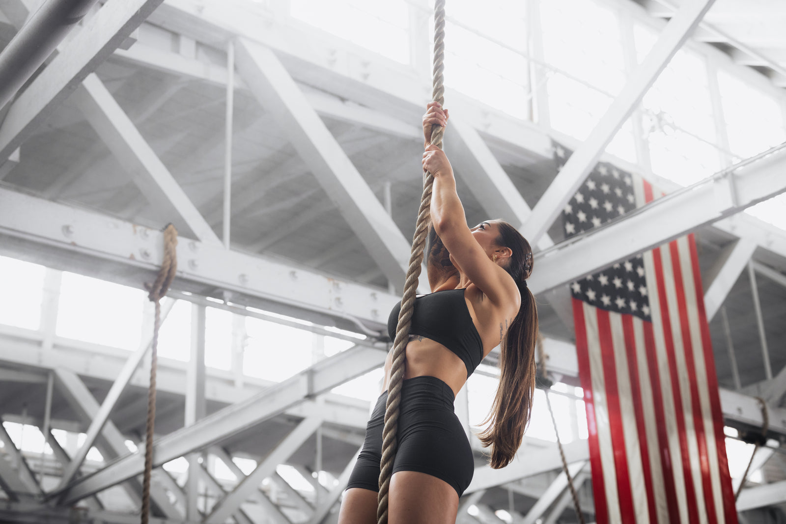 Woman climbing a rope in an industrial-style gym with an American flag hanging in the background