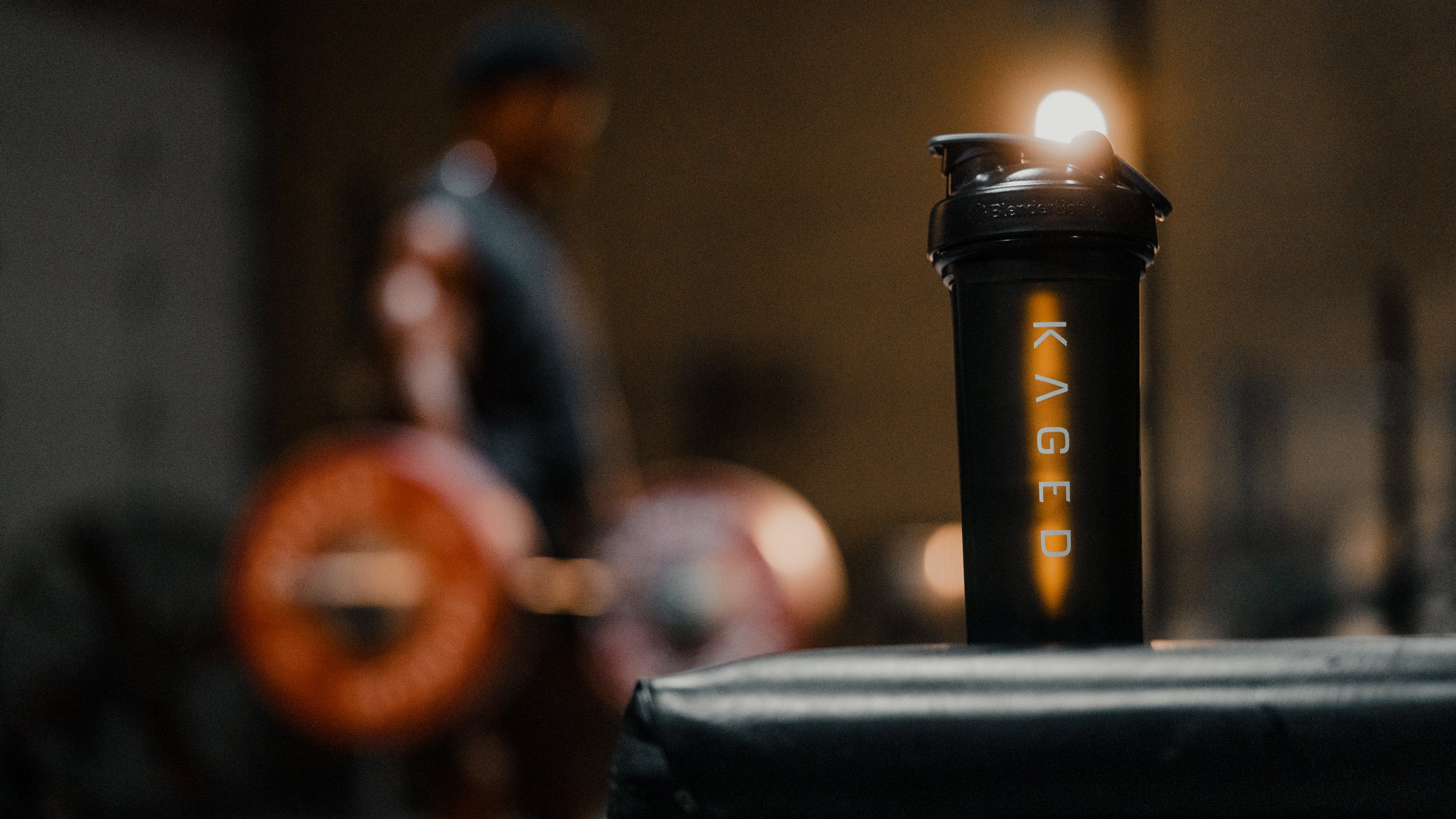 KAGED shaker bottle resting on a gym bench with a blurred weightlifter and loaded barbell in the background