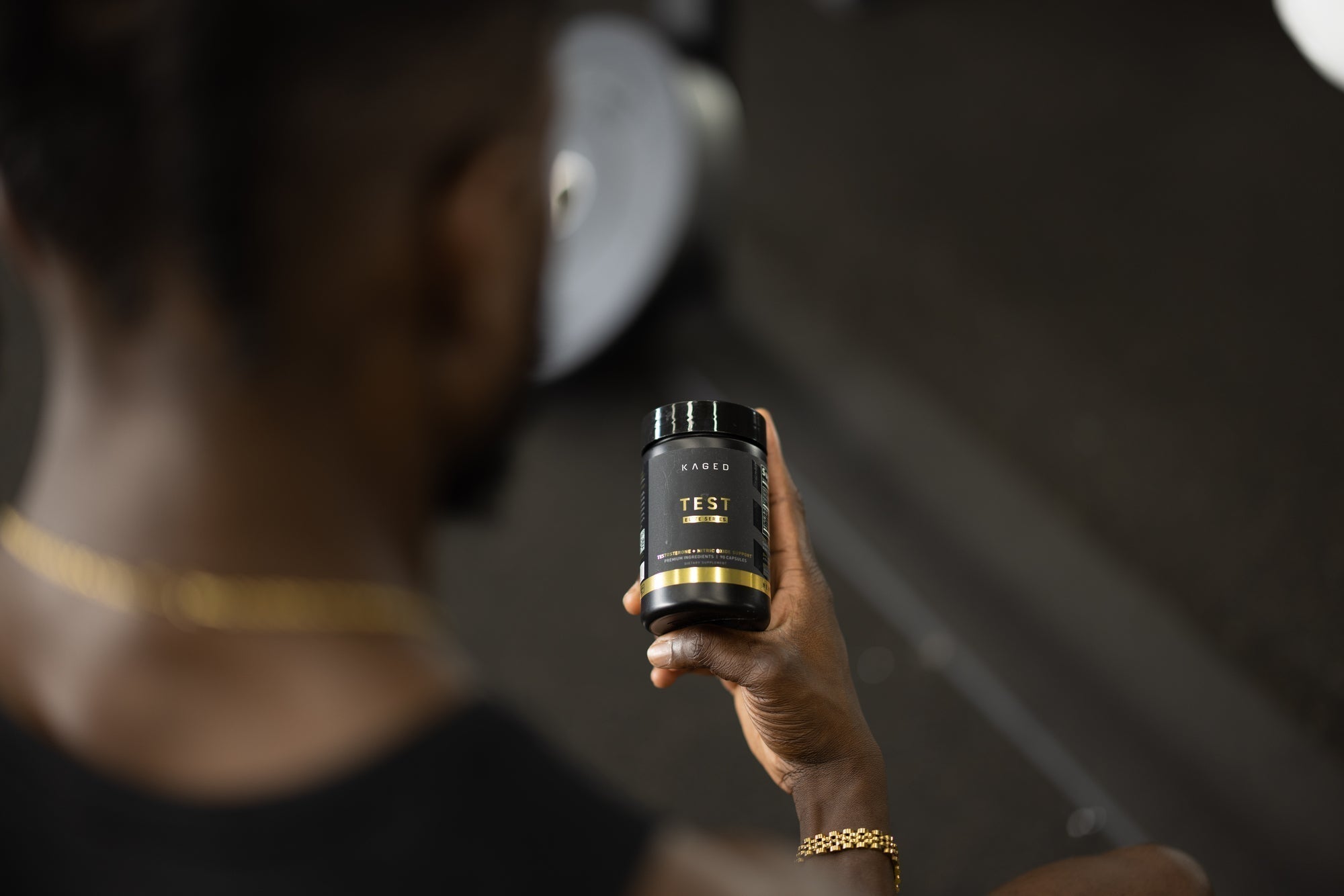 Man holding a KAGED supplement jar labeled TEST in a gym setting, hand and bottle in focus