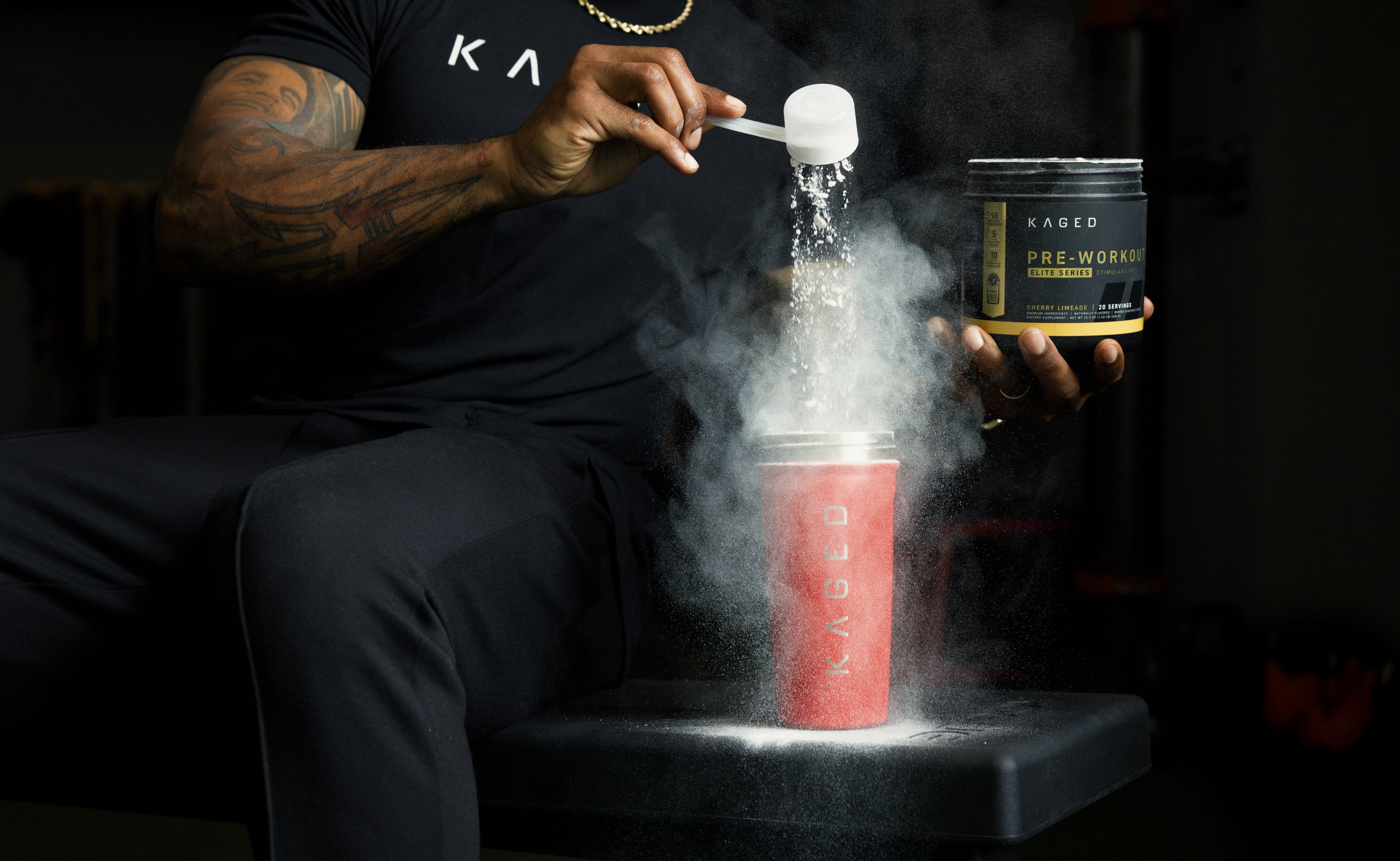 Athlete pouring KAGED pre-workout powder from a tub into a red KAGED shaker, creating a cloud of powder in a dark gym setting