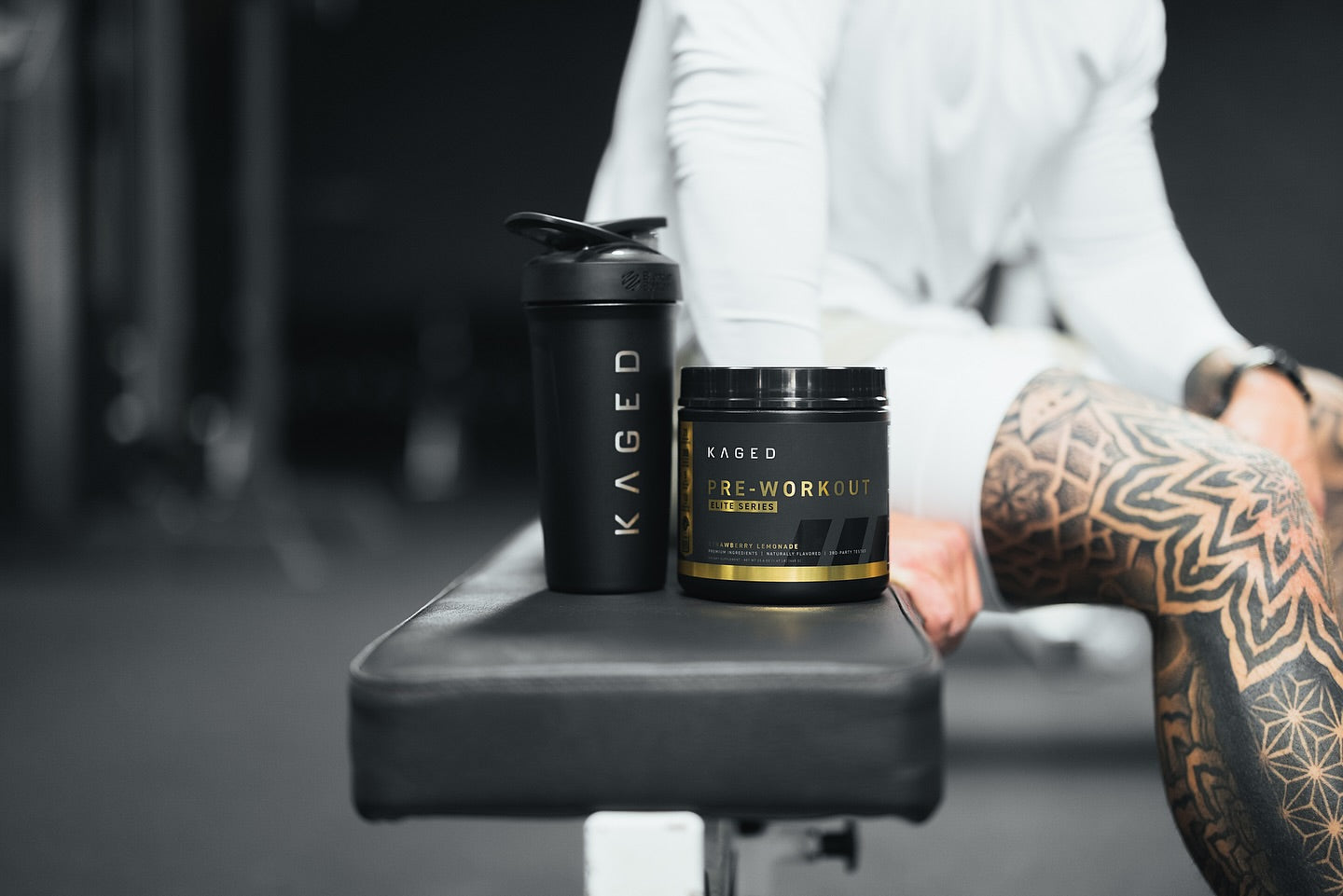Kaged pre-workout protein and shaker on a gym bench beside a tattooed athlete, ready to begin. 