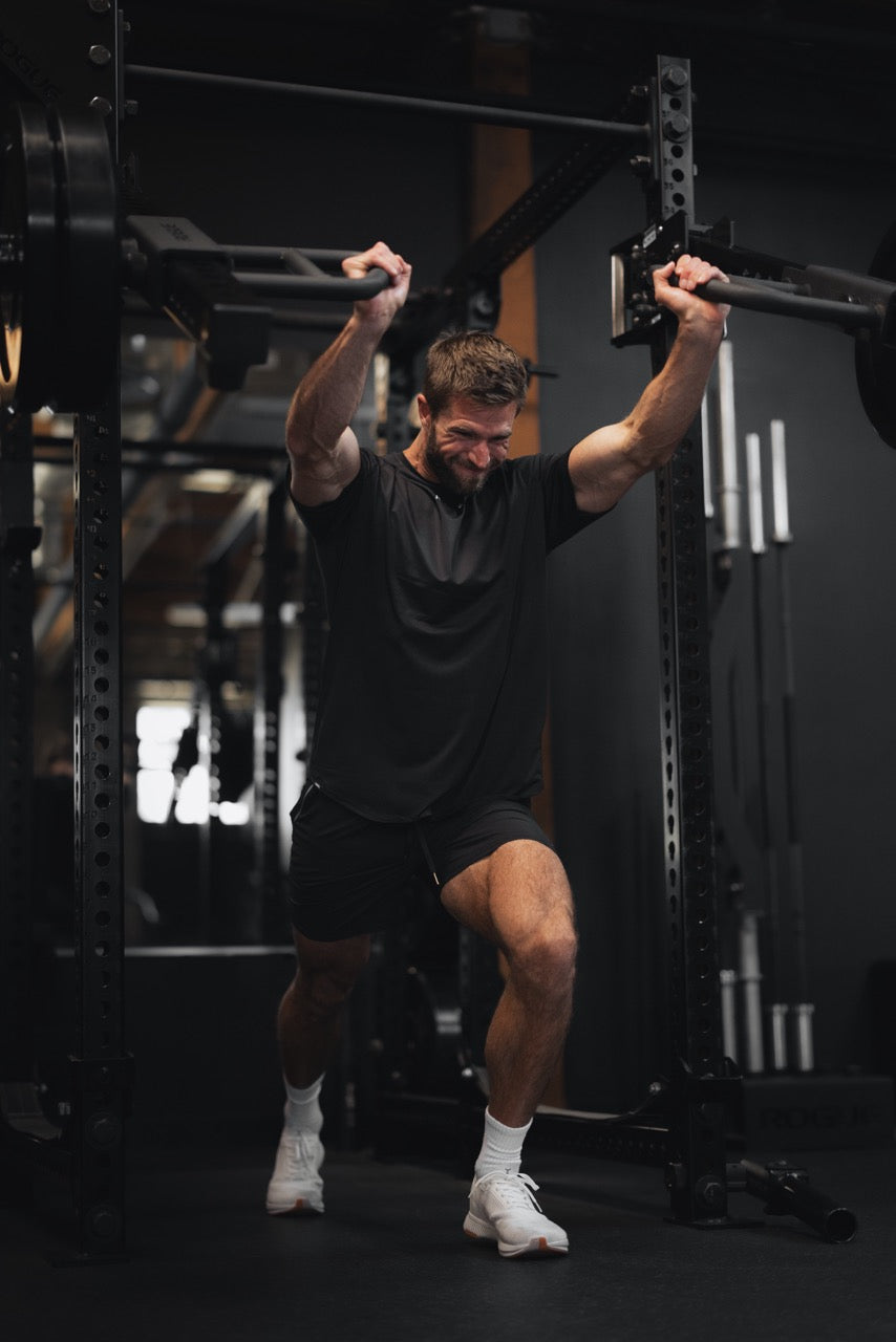 Man performing an overhead lunge inside a gym rack, holding a barbell overhead while stepping forward