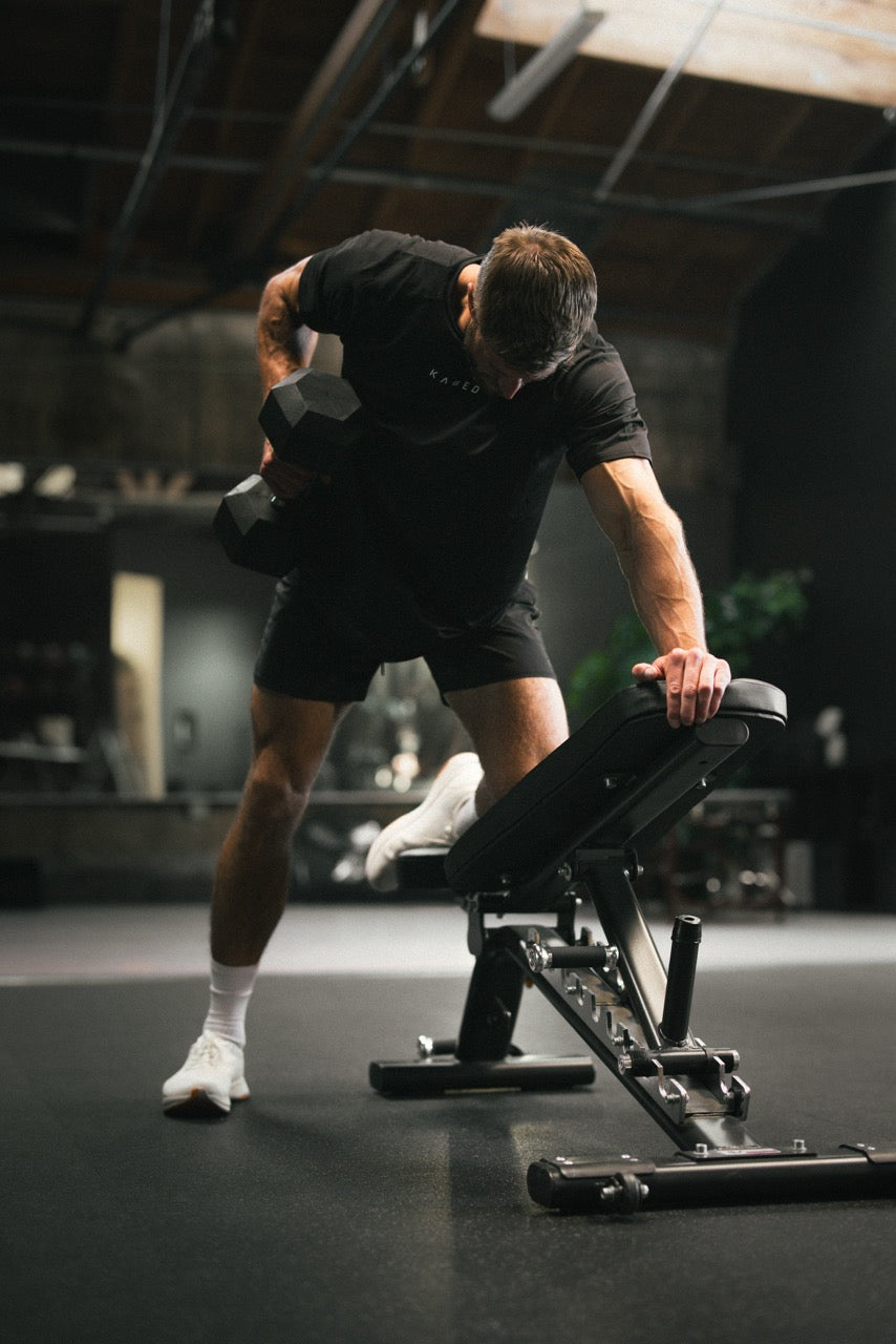 Man doing one-arm dumbbell row on an adjustable bench in a dimly lit gym, wearing a black KAGED shirt