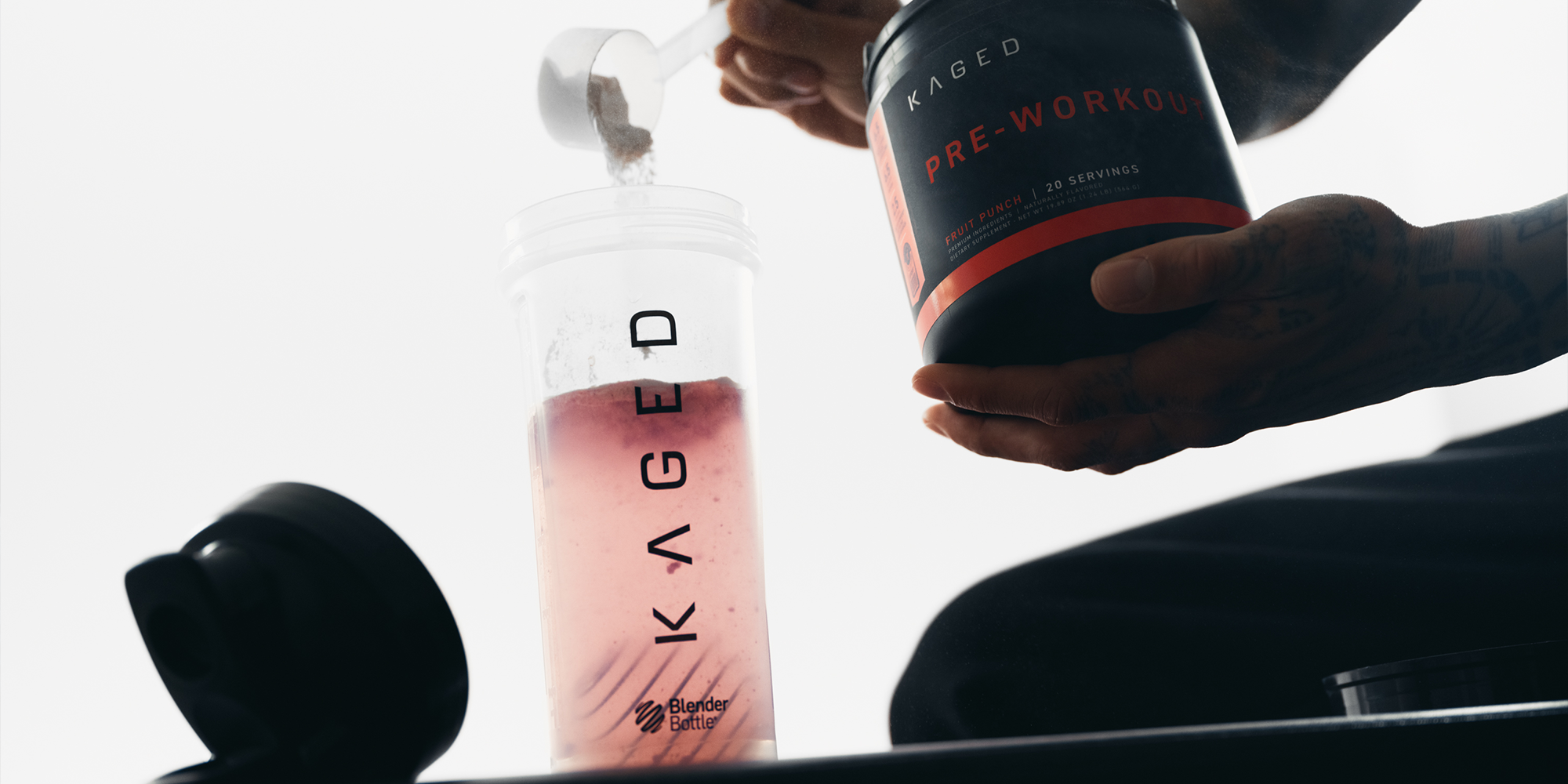 Angled view of clear Blender Bottle showcasing pink shake with hands pouring KAGED pre-workout against white background