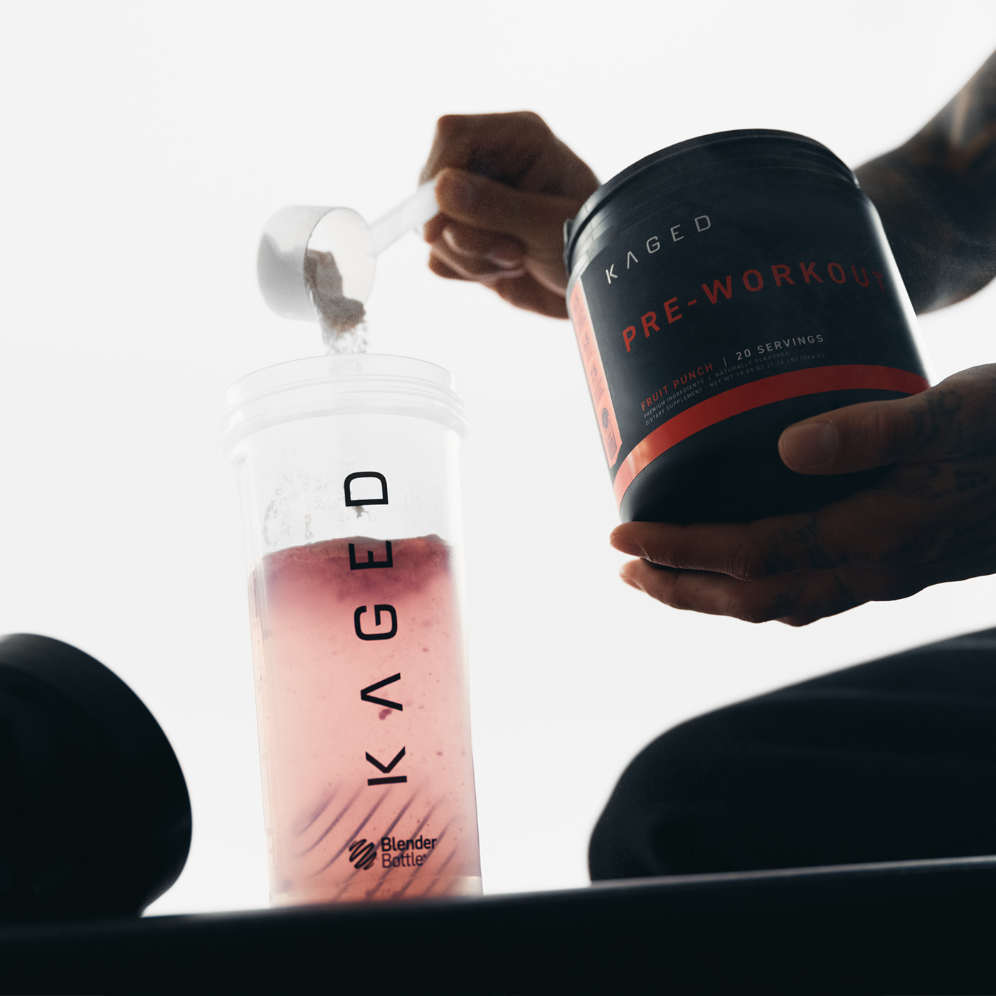 Three-quarter view of clear Blender Bottle showing KAGED logo and rounded base with pink pre-workout being poured