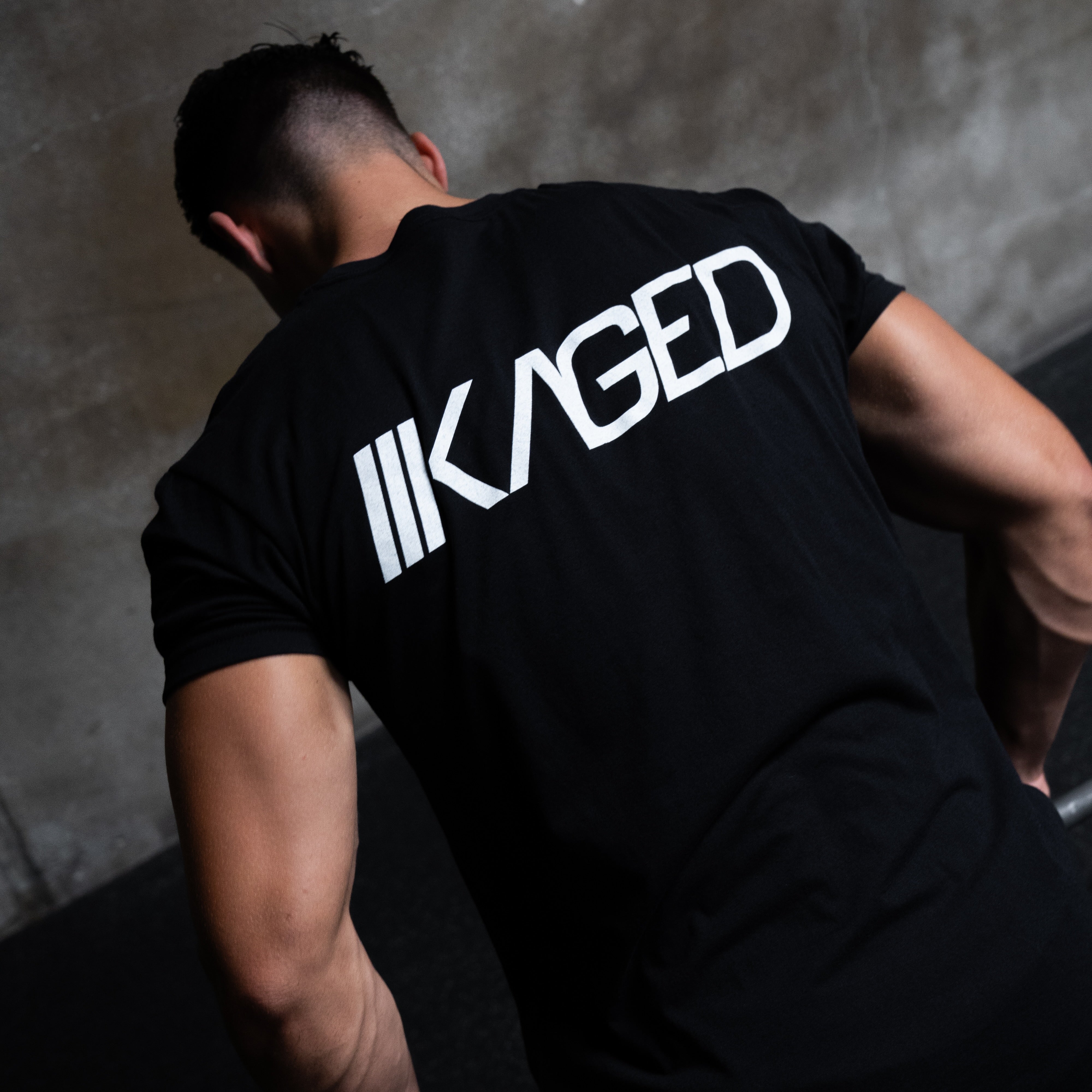 Rear view of Slant Tee in black on model showcasing large white KAGED logo across the back with concrete studio background