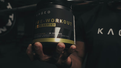 Hand holding a KAGED pre workout tub in a low-light gym setting, moody close-up showing Pre-Workout label